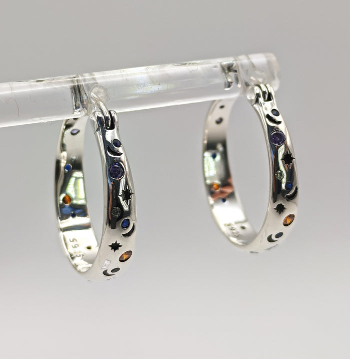 A pair of polished Sterling Silver small hoop earrings featuring an embossed or engraved pattern of crescent moons and stars, with small multi-color CZ accents.