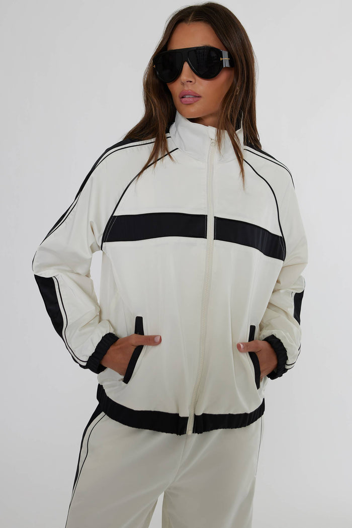 A woman modeling a white track jacket with bold black horizontal stripes and black trim, paired with matching white track pants against a plain light gray studio background.