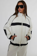 A woman modeling a white track jacket with bold black horizontal stripes and black trim, paired with matching white track pants against a plain light gray studio background.