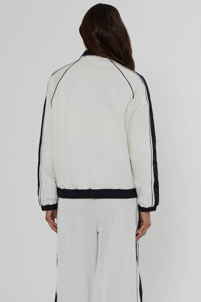 A model viewed from the back wearing a white cropped track jacket with black contrast piping and black side stripes, paired with matching white track pants against a light gray studio background.