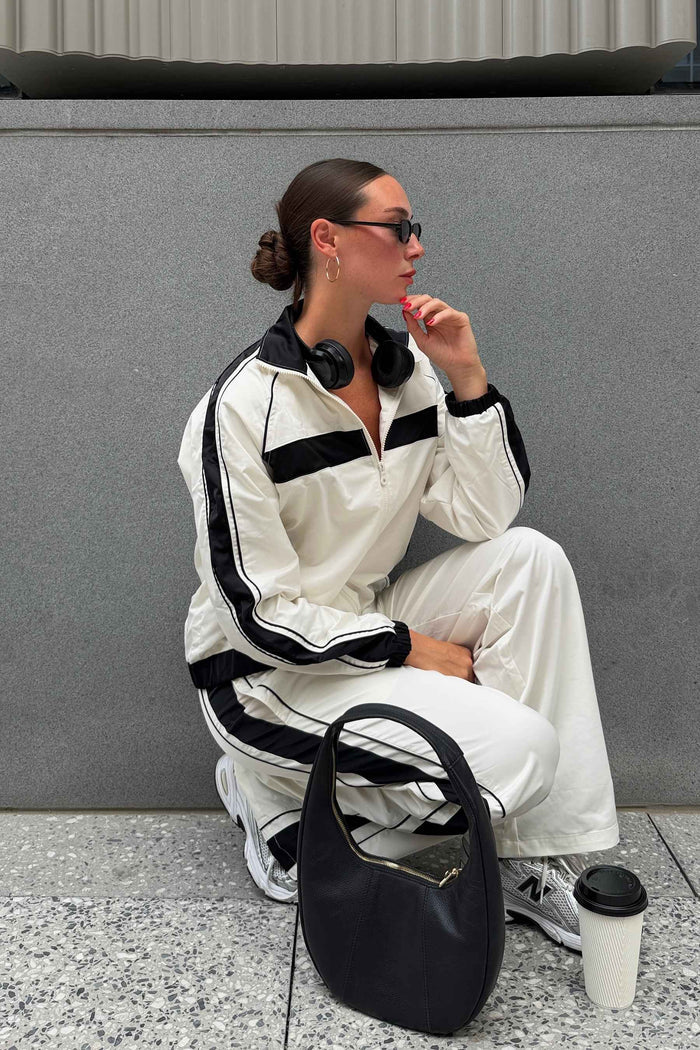 A woman wearing a matching off-white and black athletic tracksuit sits on a gray stone ledge against a gray concrete wall. She is wearing black headphones around her neck, sunglasses, and has a black handbag and a coffee cup in front of her.