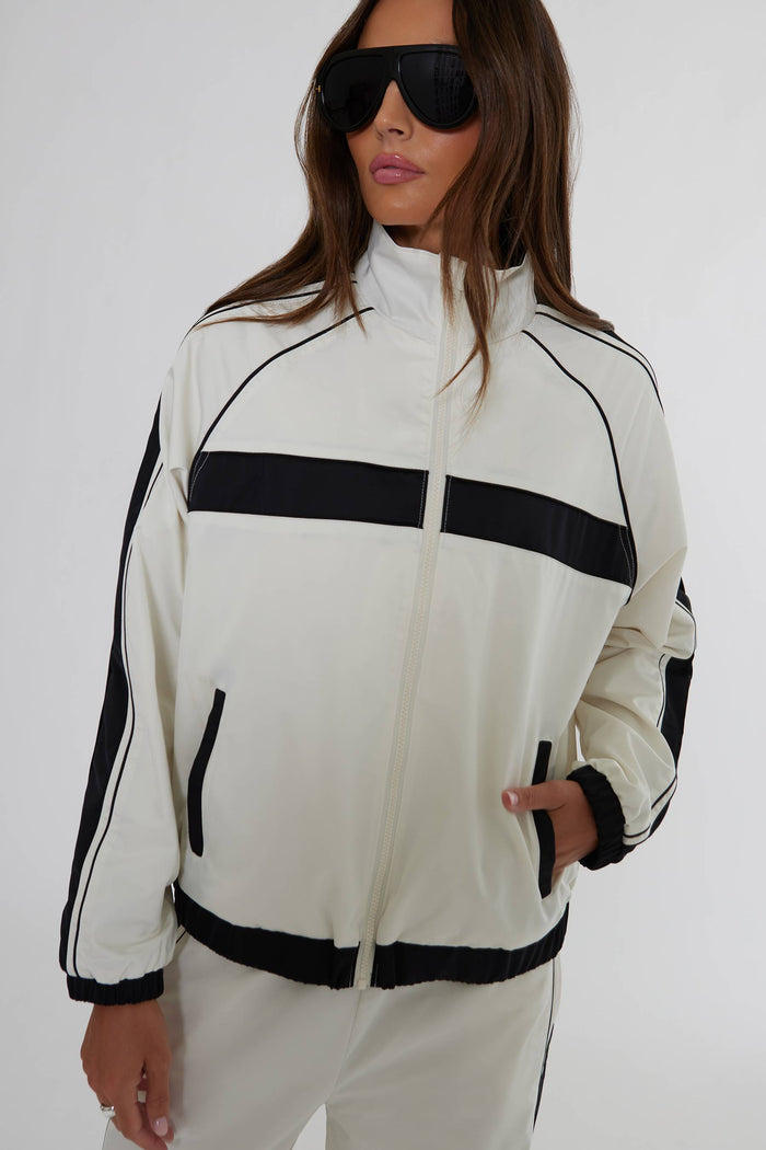 A woman wearing a cream and black track jacket with black side stripes and a black horizontal chest stripe, paired with matching cream track pants and black sunglasses, against a plain light gray background.