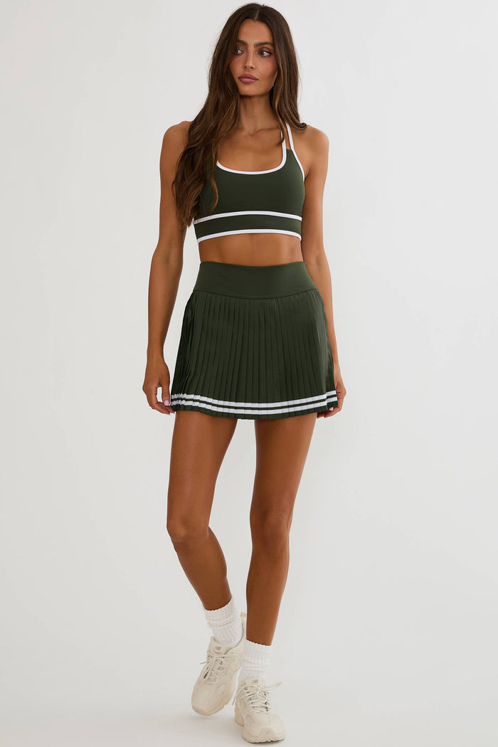 A woman modeling a matching dark green athletic set consisting of a sports bra and a pleated mini skirt, both with white trim, paired with white socks and sneakers against a plain light gray background.