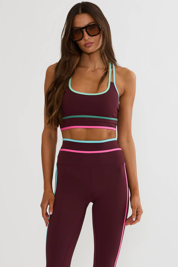 A woman modeling a matching athletic set consisting of a scoop-neck sports bra and high-waisted leggings. The activewear is primarily deep burgundy with contrasting light blue and bright pink trim details.