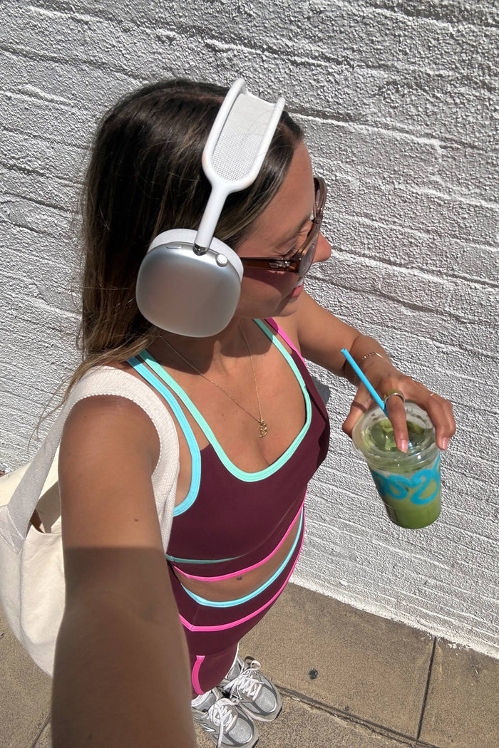 A woman wearing a white over-ear headphone-style headband accessory, a sleeveless burgundy top with turquoise and pink stripes, and holding a green drink with a blue straw, standing against a white brick wall.