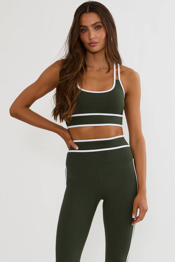 A woman modeling a matching dark green activewear set consisting of a sports bra and high-waisted leggings with white contrast trim, against a plain light gray background.