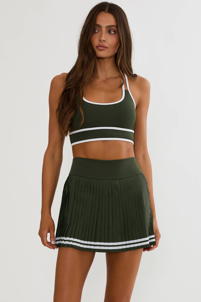 A woman modeling an olive green athletic set consisting of a sports bra and a pleated mini skirt, both featuring white trim and double white stripes near the hem, against a plain light gray background.