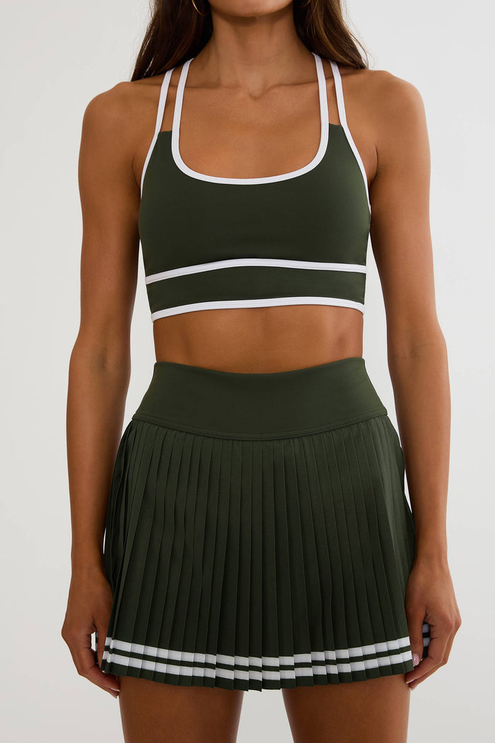 A woman wearing an olive green athletic set consisting of a sports bra and a pleated tennis skirt, both featuring white trim details, against a plain light gray background.