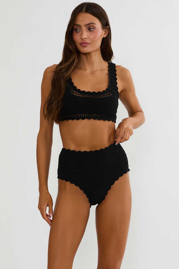Woman wearing a black crochet swimsuit on a white wall.