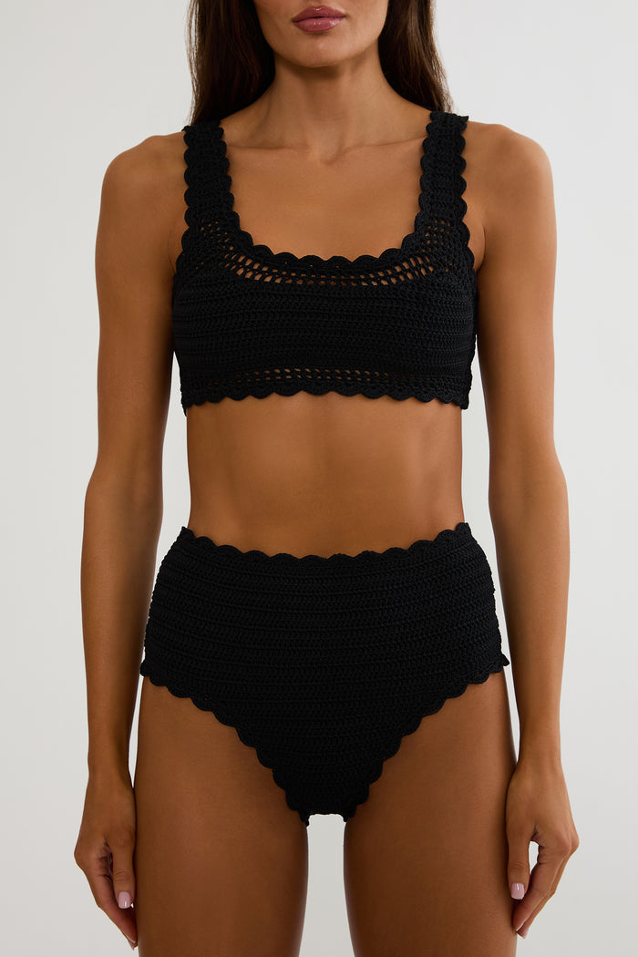 Woman wearing a black crochet swimsuit on a white wall.