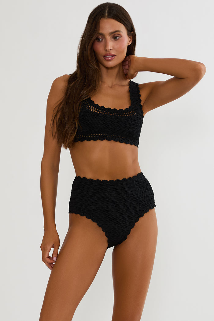 Woman wearing a black crochet swimsuit on a white wall.