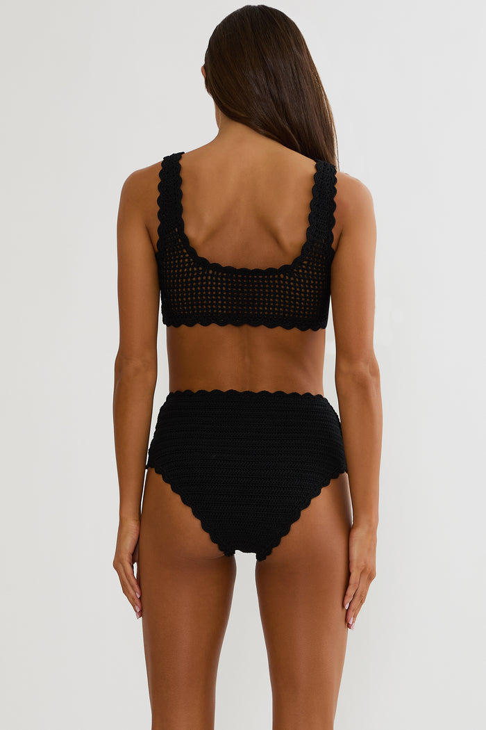 Woman wearing a black crochet swimsuit on a white wall.