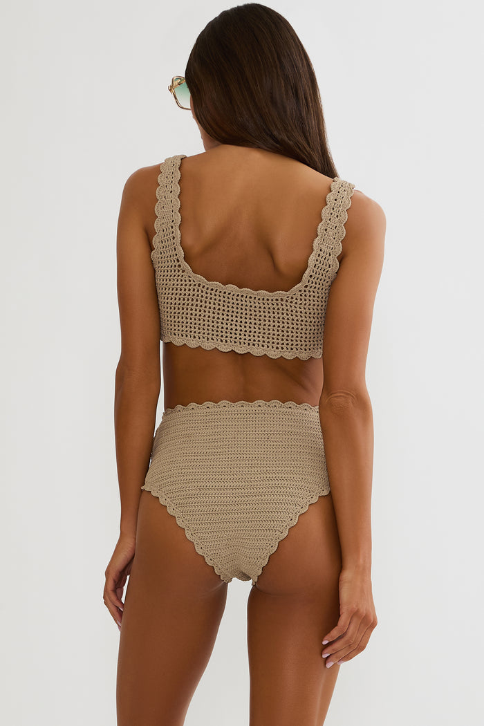 Woman wearing a tan crochet swimsuit on a white wall.