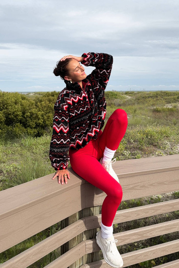 Person wearing a patterned sweater and red leggings sitting on a wooden bench with a scenic background.