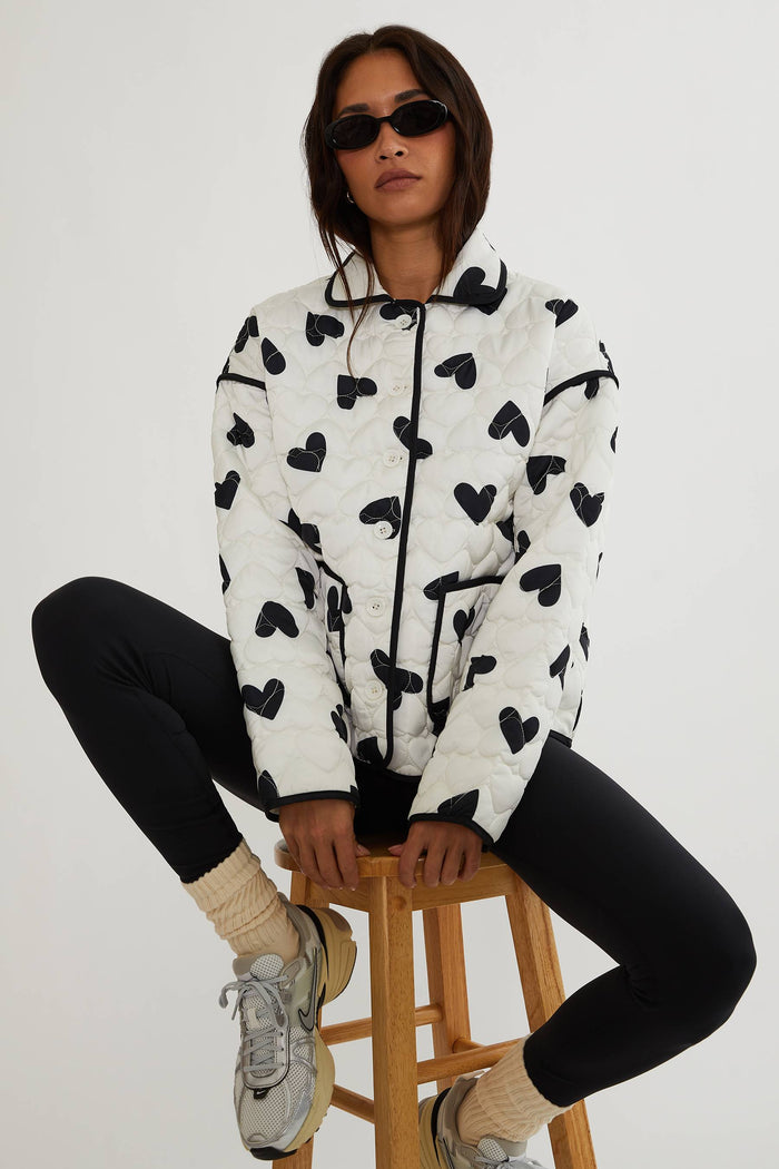 Person wearing a white hoodie with black heart patterns, sitting on a wooden stool against a white background.