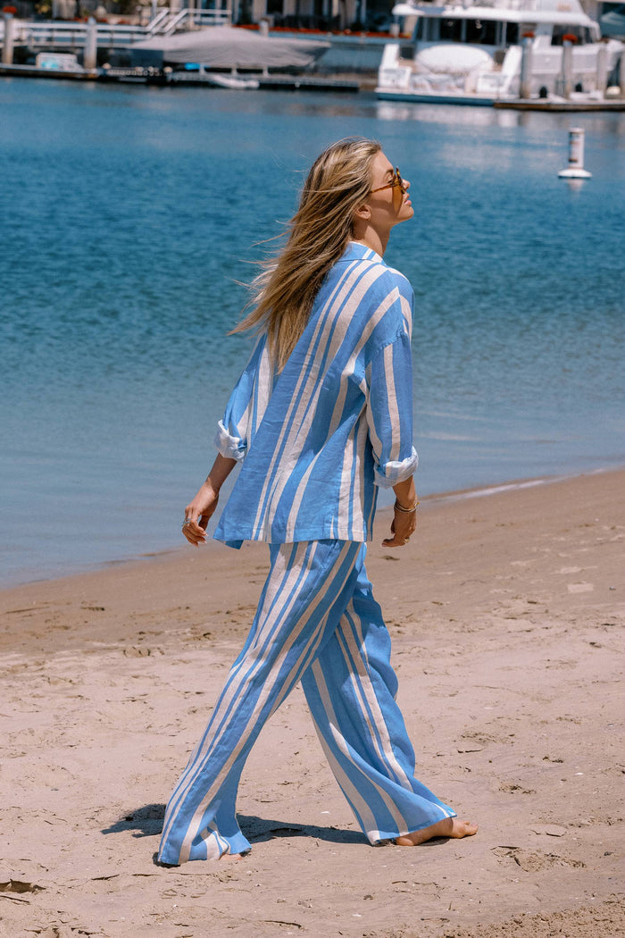 Woman in a blue and white striped outfit walking on a sandy beach with water and boats in the background.