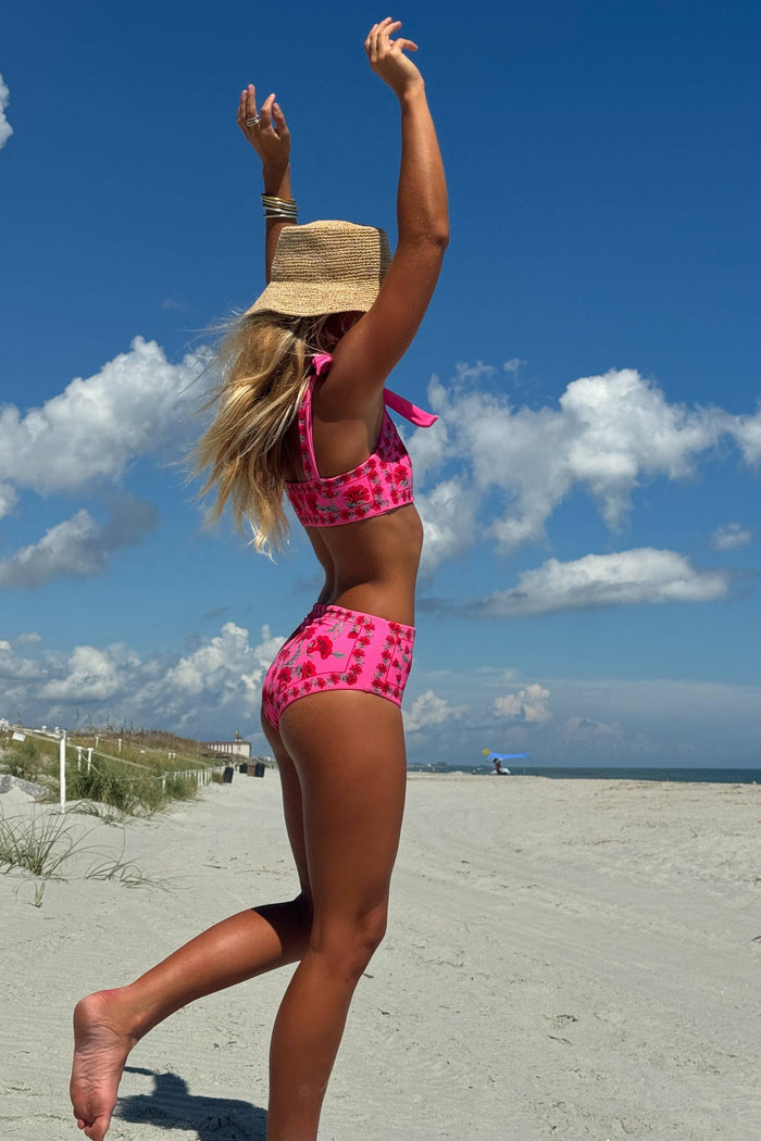 Woman in a pink bikini and straw hat on a sandy beach with blue sky and clouds.
