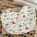 Flatlay of a handmade baby bib with a farm-themed pattern. The design includes cows, sheep, horse, chickens, blue and green tractors, red barns, and small trees on a light cream base. The bib has a cotton front and a soft terry towelling back for absorbency.
