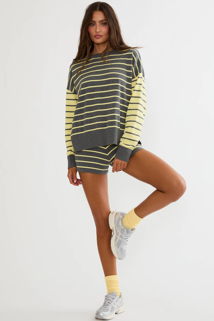 A woman modeling a matching two-piece lounge set consisting of a long-sleeve crewneck sweatshirt and shorts, both featuring dark gray and pale yellow horizontal stripes. She is also wearing light yellow socks and white sneakers against a plain light gray studio background.