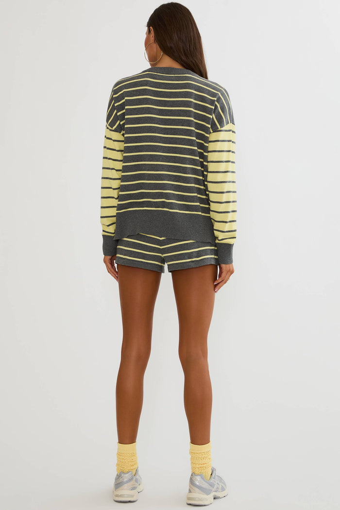 A woman shown from the back wearing a dark gray and yellow striped long-sleeve top and matching striped shorts, paired with yellow socks and gray sneakers, against a light gray studio background.