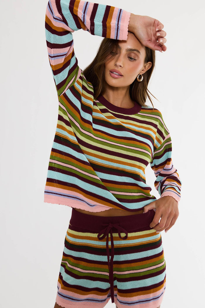 A woman modeling a matching two-piece knit lounge set with horizontal multicolor stripes in pink, orange, yellow, green, blue, and dark brown, featuring a crewneck top and drawstring shorts against a plain white background.