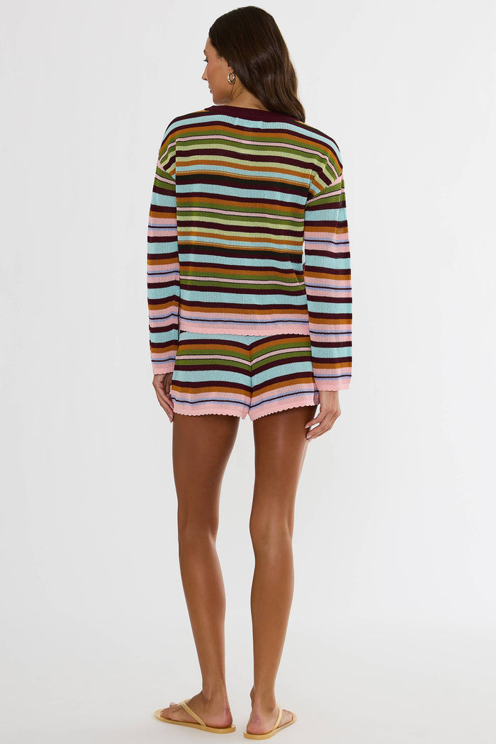 A woman viewed from the back wearing a long-sleeve knit top with horizontal multicolor stripes in pink, orange, yellow, green, blue, and brown, paired with tan flat sandals against a plain white studio background.