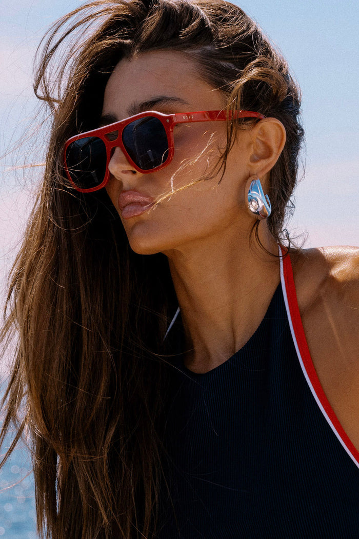 Woman wearing red sunglasses and a black tank top with white trim against a clear blue sky.