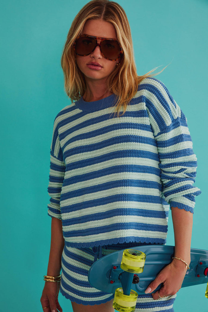 Woman wearing a blue and white striped sweater and shorts, holding a skateboard against a teal background.