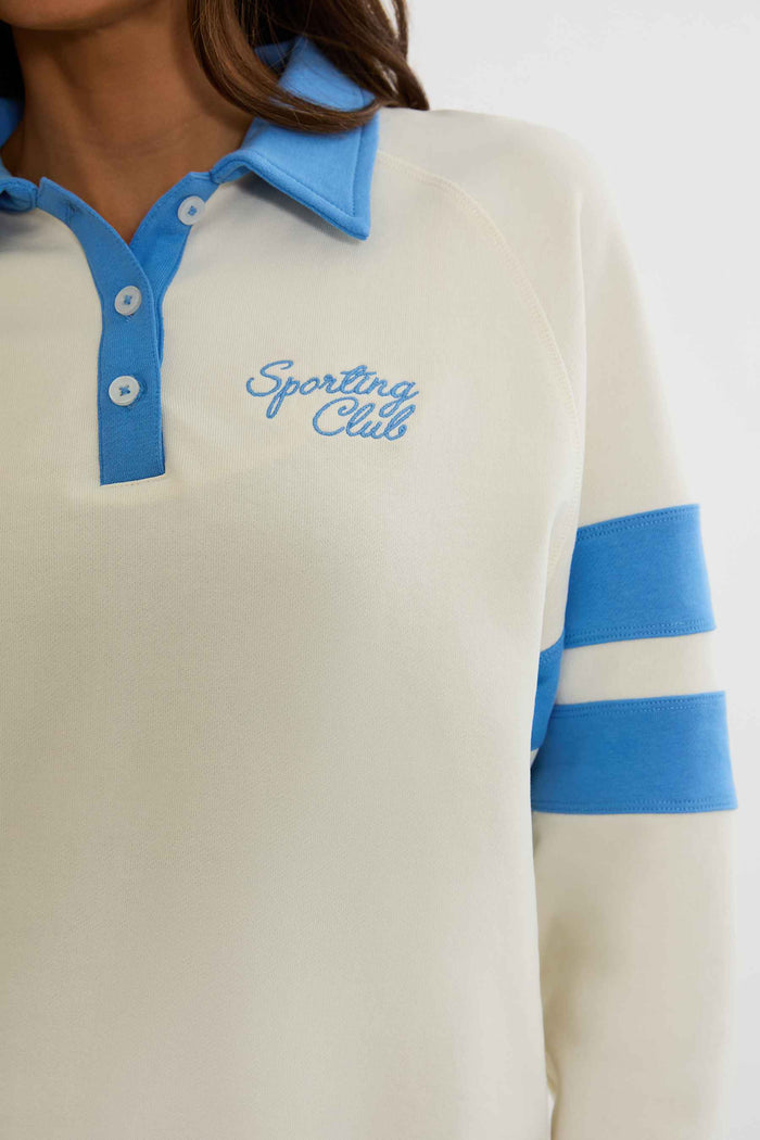 Woman wearing cream and blue polo sweater with "sports club" text on the chest.