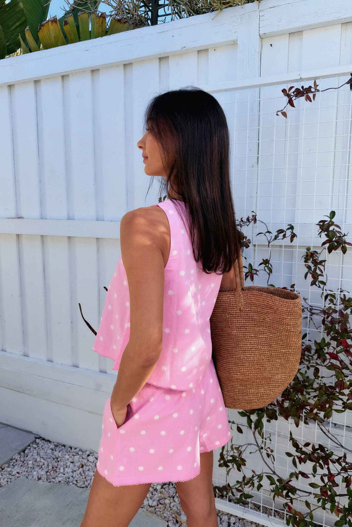 Woman in a pink polka dot dress holding a woven bag against a white fence background