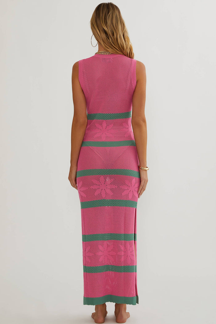 A woman viewed from the back wearing a sleeveless knit maxi dress in bright pink with horizontal teal-green stripes and floral lace-like patterning, against a plain light gray studio background.