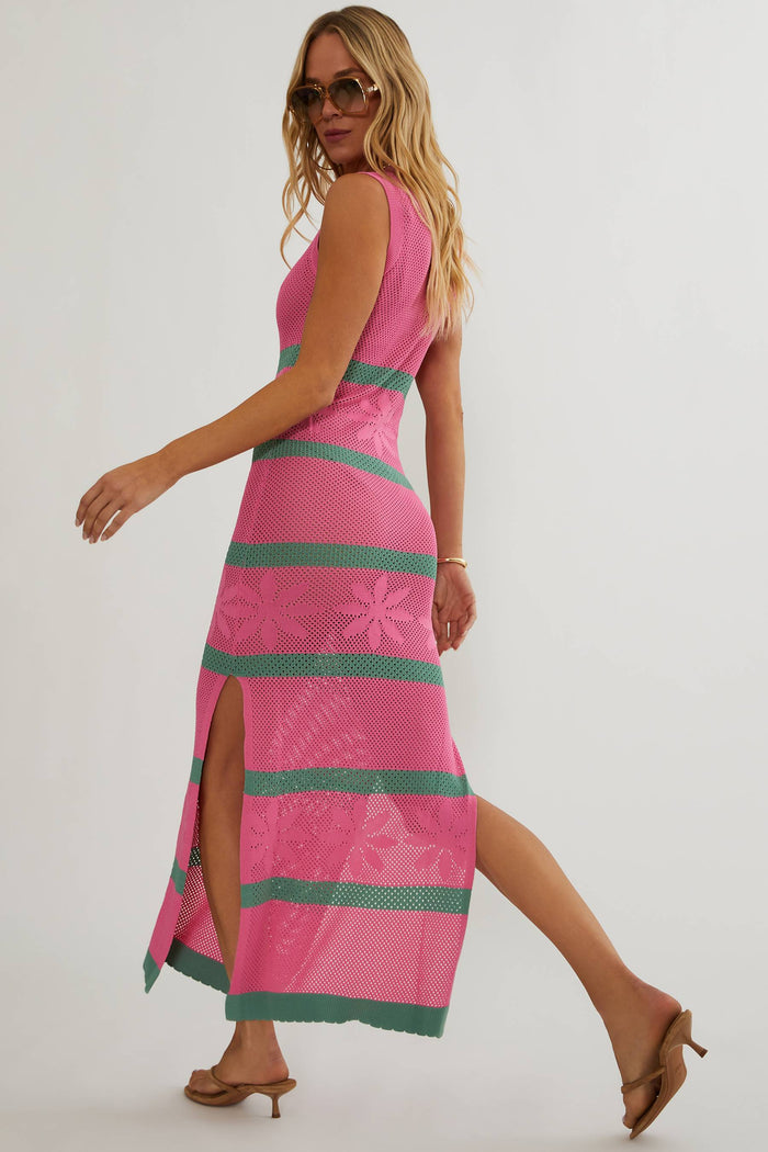 A woman modeling a sleeveless pink maxi dress with green horizontal stripes and a floral lace overlay, shown from a side-back angle against a plain light background.