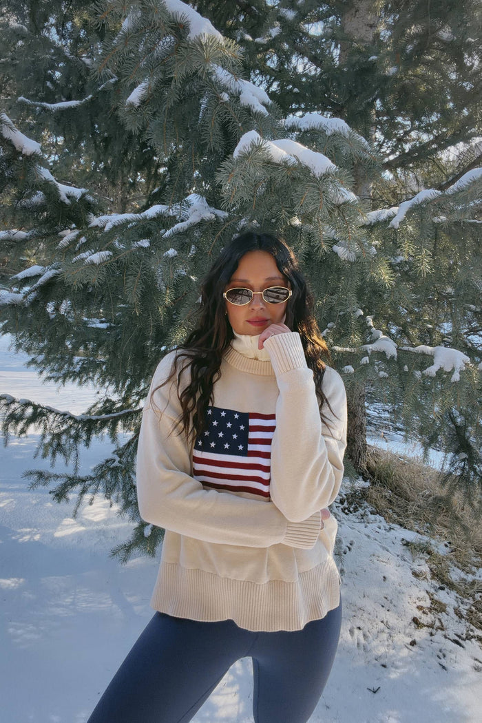 A female model wearing a white oversized crewneck sweater with a graphic of the American flag on the chest.