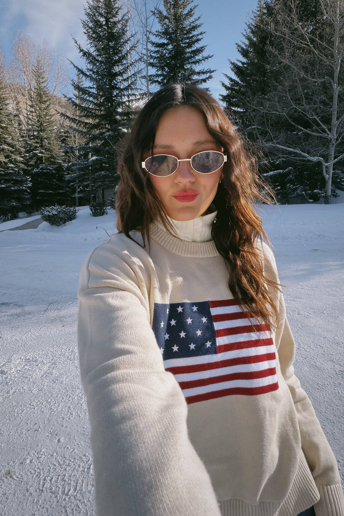 A female model wearing a white oversized crewneck sweater with a graphic of the American flag on the chest.