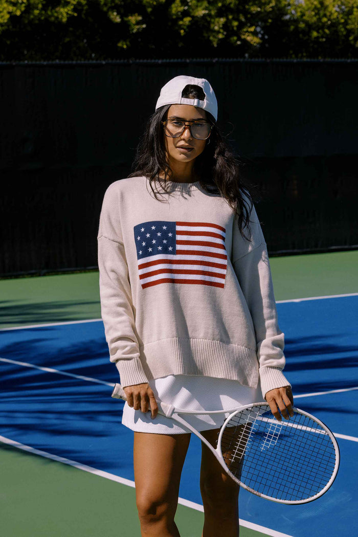 A female model wearing a white oversized crewneck sweater with a graphic of the American flag on the chest.
