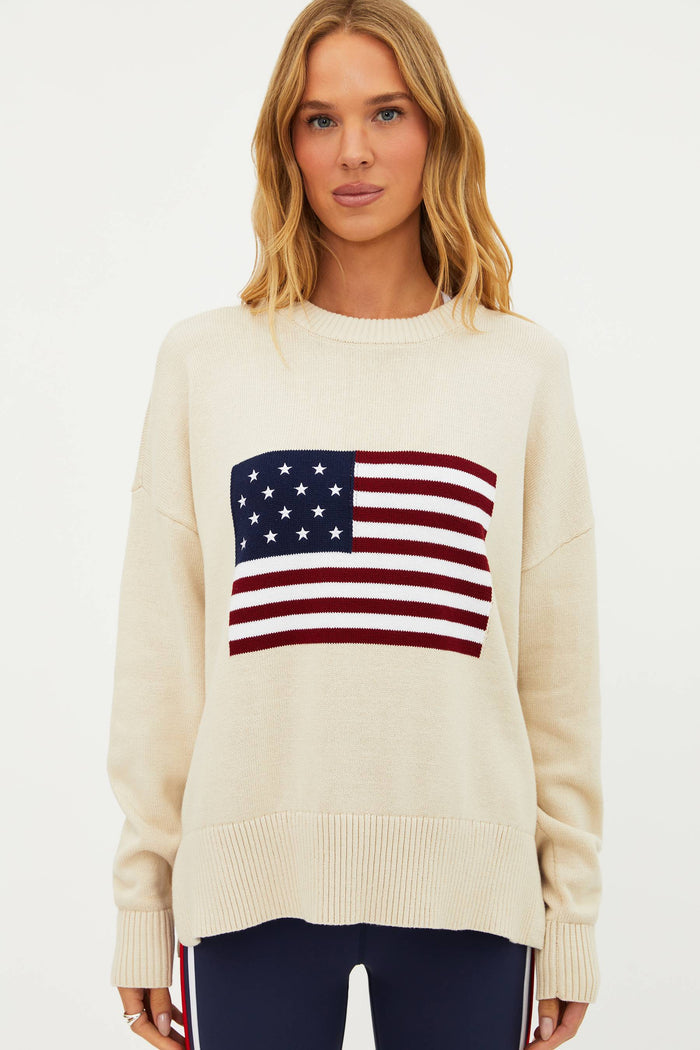 A female model wearing a white oversized crewneck sweater with a graphic of the American flag on the chest.