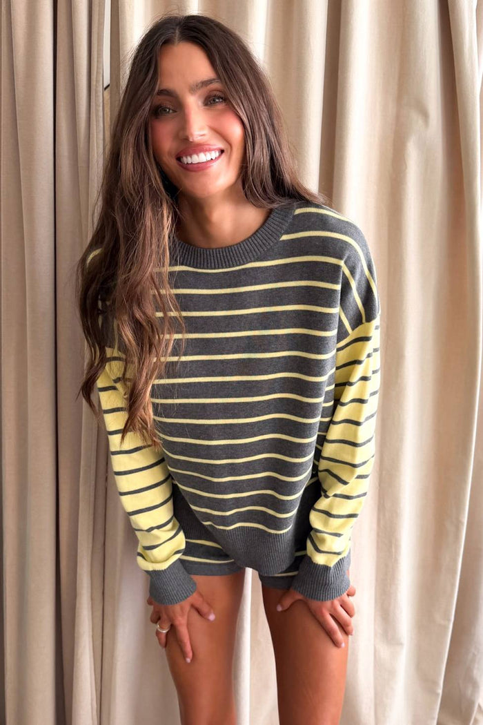 A woman smiling while wearing a gray and yellow striped knit sweater with solid yellow sleeves, standing in front of beige curtains.