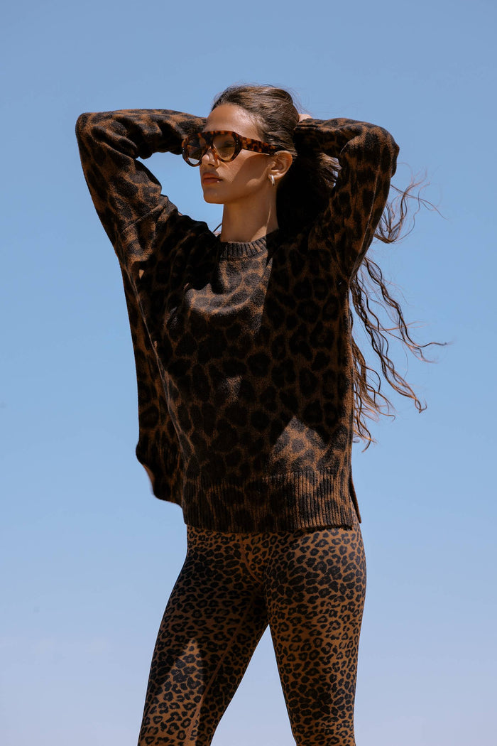 A woman wearing a brown and black leopard print knit sweater and matching leopard print leggings, posing outdoors against a clear blue sky.