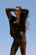 A woman wearing a brown and black leopard print knit sweater and matching leopard print leggings, posing outdoors against a clear blue sky.