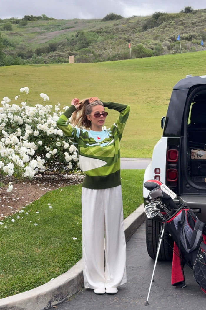 Woman in a green and white sweater and white pants standing next to a car with golf clubs on a grassy area.