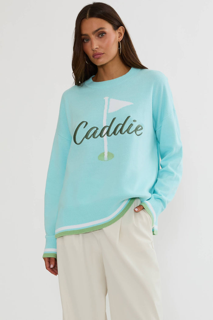 A woman modeling a light blue crewneck sweatshirt with a golf flag graphic and the word 'Caddie' across the chest, paired with cream-colored trousers, against a plain light gray background.