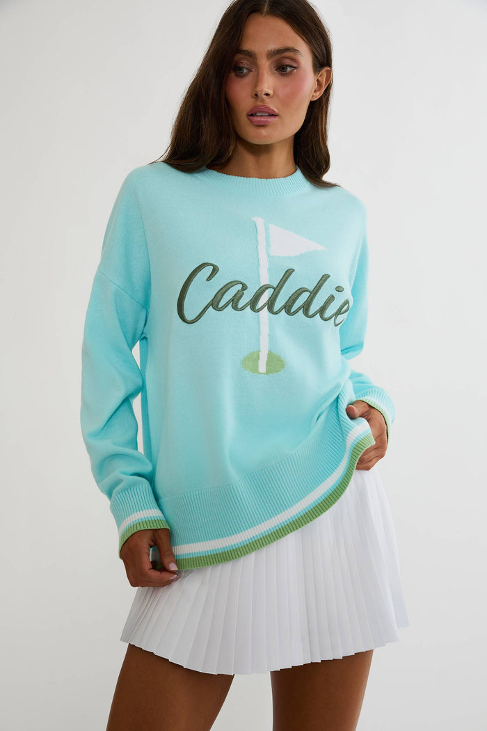 A woman modeling a light aqua long-sleeve knit sweater with 'Caddie' and a golf flag graphic on the front, paired with a white pleated tennis skirt against a plain light gray studio background.