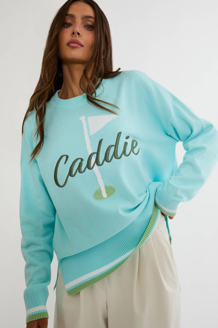 A woman wearing a light blue-green crewneck sweatshirt with a golf flag graphic and the word 'Caddie' across the front, paired with cream trousers, against a plain light gray studio background.