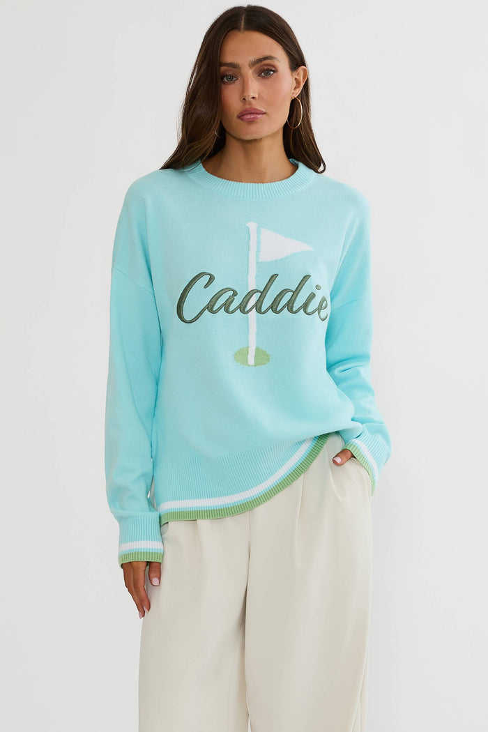 A woman modeling a light blue crewneck sweater with a green and white golf flag graphic and the word 'Caddie' across the chest, paired with cream-colored trousers, against a plain white studio background.