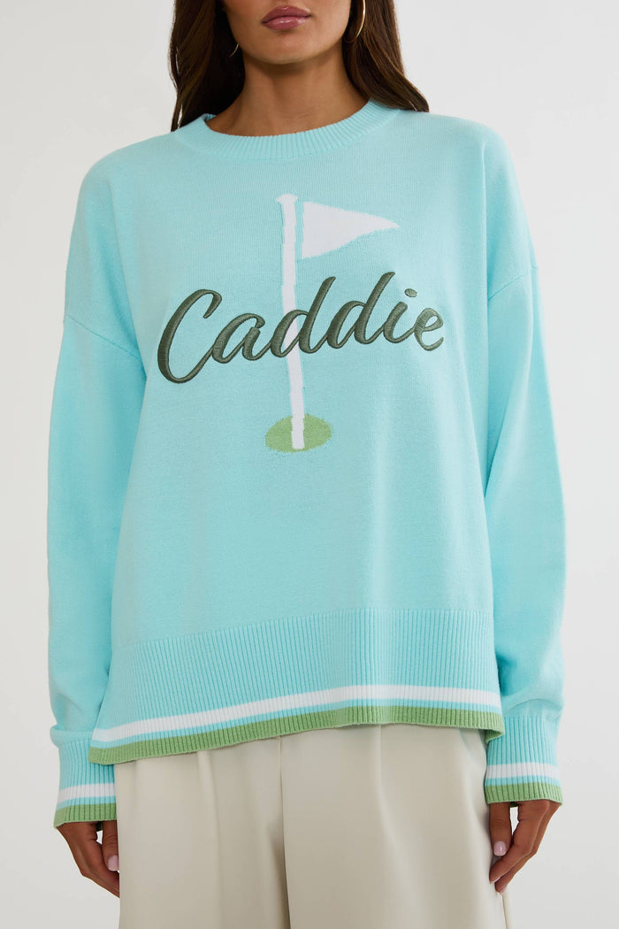 A woman wearing a light aqua long-sleeve knit sweater with a golf flag graphic and the word 'Caddie' across the chest, paired with light beige pants against a plain light background.