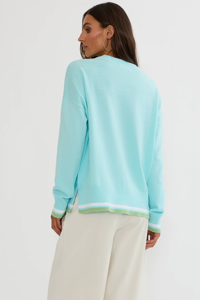 A woman shown from the back wearing a light aqua knit sweater with white and mint green striped ribbing at the hem and cuffs, paired with off-white trousers, against a plain white studio background.