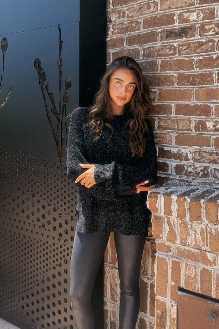 A woman with long wavy brown hair stands outdoors against a brick wall and dark metal wall, wearing a black textured knit sweater and black leggings.