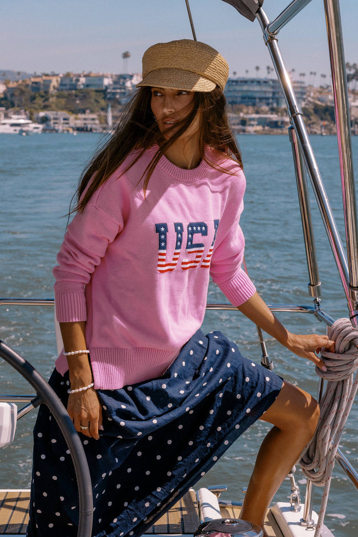 Woman on a sailboat wearing a pink sweater with 'BICIV' text and a navy skirt.