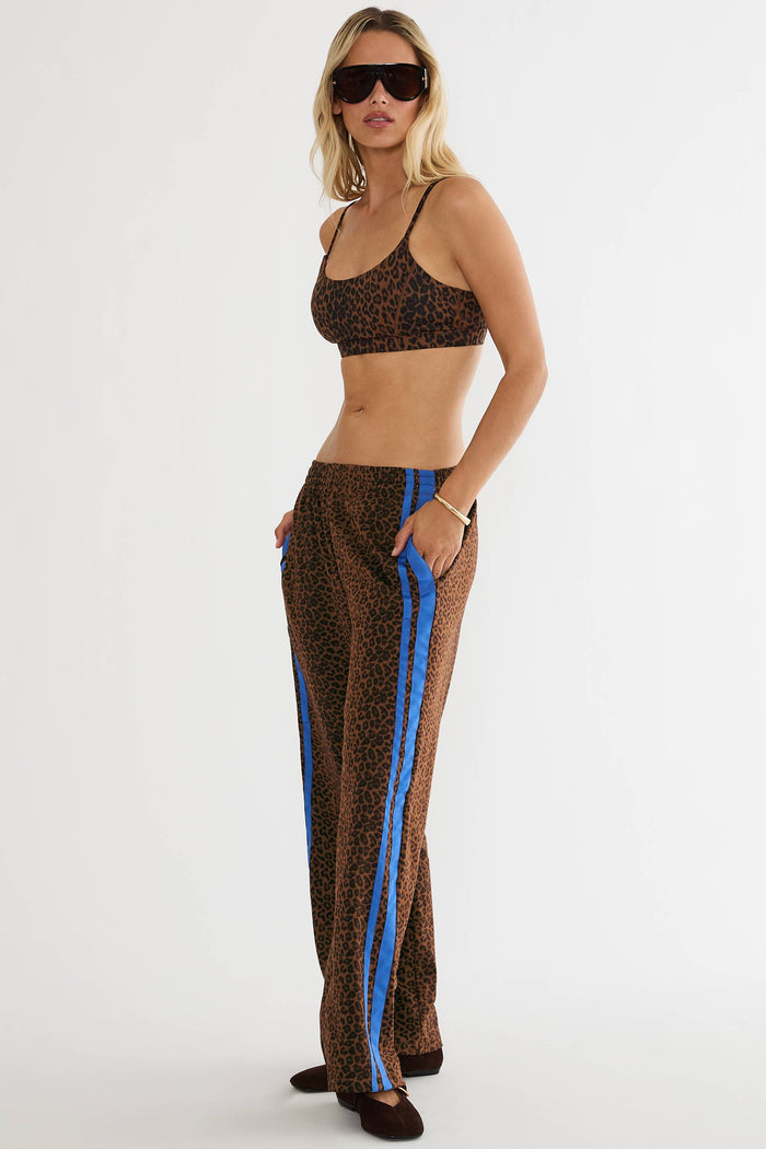 A woman modeling a matching leopard-print two-piece set consisting of a sports bra and wide-leg pants with bright blue side stripes, photographed against a plain white studio background.