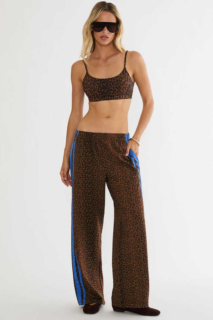 A woman modeling a matching leopard-print activewear set consisting of a sports bra and wide-leg pants with blue side stripes, photographed against a plain white studio background.